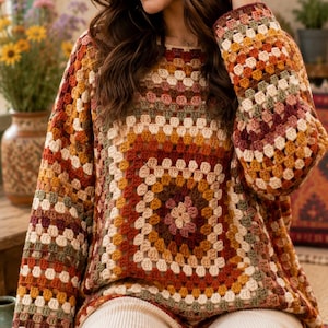 May include: A long-sleeved jumper crafted from crocheted yarn in a granny square pattern. The garment features a multi-coloured palette of earthy tones including rust, mustard yellow, cream, sage green, and deep burgundy in a repeating block design.