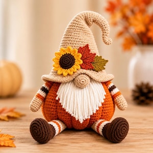 Op de afbeelding: Een gehaakt kabouterfiguur met een beige puntmuts versierd met een gele zonnebloem en twee stoffen bladeren. De kabouter heeft een ronde neus, een witte garen baard en een pompoenvormig oranje lichaam met gestreepte armen en bruine voeten.