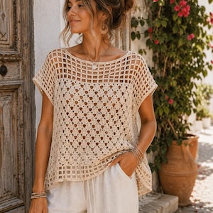 May include: A beige crochet top featuring a wide boat neckline and short sleeves. The garment displays a combination of an open grid pattern at the top and hem, with a repeating geometric diamond motif throughout the center of the bodice.