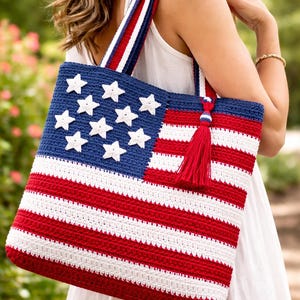 Op de afbeelding: Gehaakte draagtas met het ontwerp van de Amerikaanse vlag. De tas heeft een blauw bovenstuk met witte sterren en rood-witte strepen. De bandjes zijn rood, wit en blauw. Een rode kwast hangt aan de zijkant.