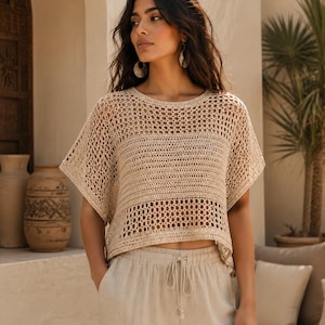 Può includere: Un top all'uncinetto beige con scollo rotondo, maniche corte e ampie e una combinazione di punti traforati e pieni. Il capo presenta una silhouette corta e squadrata ed è abbinato a pantaloni beige coordinati con coulisse in vita.