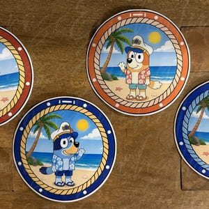 May include: Four circular coasters with cartoon dog characters in captain's hats and beachwear, set against a beach scene with palm trees and the sea. The coasters have a rope-style border in red, orange, and blue.