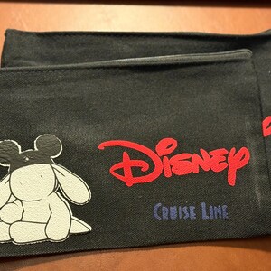 Può includere: Due pochette con cerniera in tela nera. Una pochette presenta un personaggio dei cartoni animati in bianco e nero e il logo Disney rosso con "Cruise Line" in testo blu. La seconda pochette è parzialmente visibile dietro la prima.