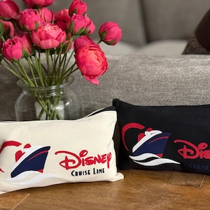Op de afbeelding: Twee rechthoekige Disney Cruise Line etuis met ritssluiting. Eén is gebroken wit, de andere zwart. Beide hebben een rood, wit en blauw cruiseschip-ontwerp en het Disney Cruise Line-logo in rood. Een vaas met roze bloemen staat achter de etuis.