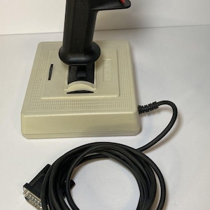 May include: A vintage beige joystick controller featuring a black handle with a red top button and a red trigger. The base is square with a textured surface and a black cable attached, ending in a multi-pin connector plug for gaming consoles.