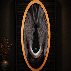 Op de afbeelding: Een modern wandkunstwerk met een ovaal ontwerp van zwart en grijs draad. Het ovaal is omlijst met een gouden rand en geplaatst tegen een donkere, getextureerde achtergrond. Een kleine groene bol hangt in het midden.
