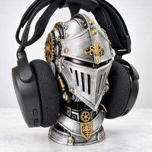 Op de afbeelding: Een zwarte over-ear hoofdtelefoon rustend op een zilveren en gouden ridderhelm-buste. De helm heeft mechanische tandwieldetails, gouden fleur-de-lis accenten en een vizier. De hoofdtelefoon heeft een brede hoofdband en zwarte oorkussens over de helm.
