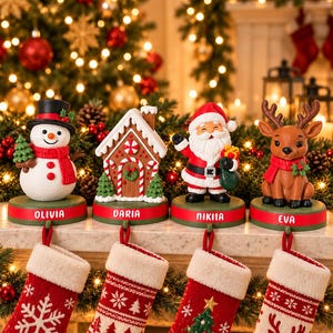 May include: Four Christmas stocking holders featuring a snowman, a gingerbread house, Santa Claus, and a reindeer. Each figure sits on a green base with a red stripe and the names OLIVIA, DARIA, NIKITA, and EVA. Four red stockings hang below.