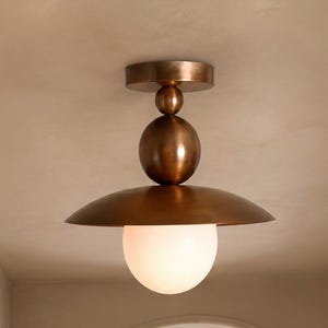 May include: A flush mount ceiling light fixture featuring a metallic bronze finish with a central spherical accent and a wide, shallow disc-shaped shade. A single white frosted glass globe bulb hangs downward from the center of the metal shade.