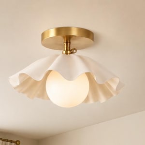 May include: A flush mount ceiling light featuring a brass finish circular canopy and a central stem. The fixture includes a white, ruffled, scalloped-edge shade surrounding a spherical, frosted glass globe bulb that emits a soft, warm light.