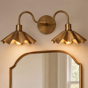 May include: A bathroom vanity with a gold-toned mirror, faucet, and sconces. The sconces have ruffled shades. A vase with greenery and a soap dish are on the marble countertop. A towel hangs on a gold towel ring.