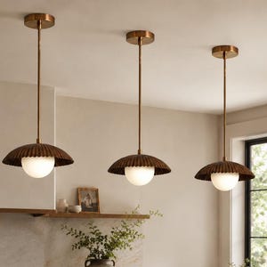 May include: Three pendant lights with a bronze finish hang above a kitchen island. Each light features a scalloped shade and a spherical, frosted glass globe. The kitchen has a marble countertop and wooden cabinetry.