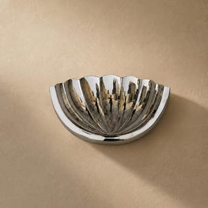 May include: A polished silver-toned bowl shaped like a seashell with deep, vertical fluted ridges radiating from the centre. The metallic surface has a high-gloss finish that reflects the surrounding environment, creating a bright, reflective appearance.