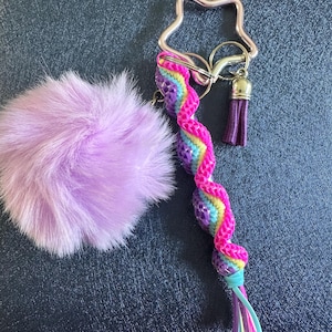 May include: A keychain featuring a star-shaped clasp, a fluffy lavender pom-pom, and a rainbow-coloured braided charm. The charm has a purple tassel and a turquoise, pink, and yellow tail. The keychain is on a dark blue surface.