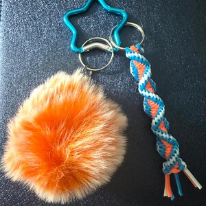 May include: A keychain with an orange fluffy pom-pom, a blue star-shaped clasp, and a braided blue, white, and orange cord. The keychain has silver metal rings. The pom-pom is made of soft, textured material.