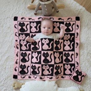 Op de afbeelding: Een baby ligt op een roze en zwarte deken met kattenpatroon, met een bijpassend kussen. Een bruin konijnenknuffel en een wit knuffeldier zijn ook in de afbeelding te zien. De deken heeft een dambordpatroon met afwisselende zwarte en roze katten silhouetten.