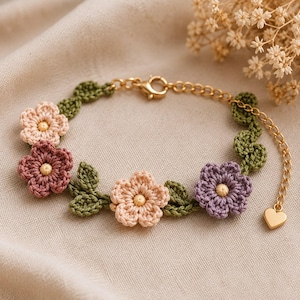 May include: A crochet bracelet featuring small flowers in shades of light pink, dusty rose, and lavender, interspersed with green crocheted leaves. The bracelet includes a gold-toned metal chain, a lobster clasp, and a small heart-shaped charm.
