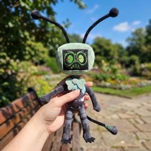 May include: A plush toy robot with a green head, black antennae, and embroidered facial features. The robot has a gray body, arms, and legs, with a tail. The toy is held against a blurred background of a park.