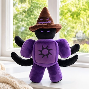 Puede incluir: Un peluche con cabeza negra y ojos morados brillantes, que lleva un sombrero puntiagudo marrón con un diseño de boca morada dentada. El cuerpo es morado con un emblema negro en forma de sol en el pecho y cuatro extremidades negras a los lados.