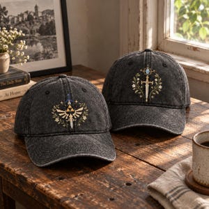 Könnte beinhalten: Zwei anthrazitfarbene Baseballkappen im Acid-Wash-Look. Die linke Kappe zeigt ein gesticktes goldenes Triforce-Emblem mit Schwert und Blumenkranz. Die rechte Kappe zeigt ein gesticktes silbernes Schwert, umgeben von einem Blumenkranz und einem blauen Edelstein.
