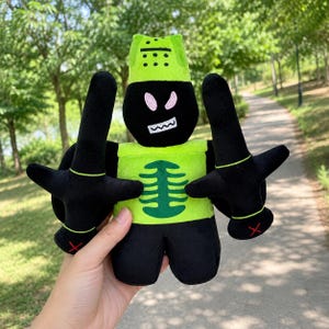 Puede incluir: Un peluche con forma de caballero en tela negra y verde lima. La figura presenta un casco verde lima, una cara negra con ojos rosas y una boca blanca, una pechera verde lima con un gráfico de caja torácica verde oscuro y dos espadas negras.