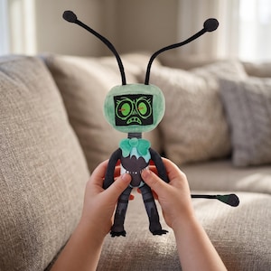 May include: A plush toy robot with a green head and a black body. The robot has two black antennae and a green bow tie. The robot's face has two glowing green eyes and a frowning mouth. The robot is being held by a person.