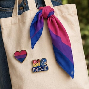 May include: A cream-colored tote bag with a heart-shaped pin and a pin that says "BI & PROUD". A scarf tied to the handle displays the bisexual pride flag colors: pink, purple, and blue. The bag is carried by a person wearing blue jeans.