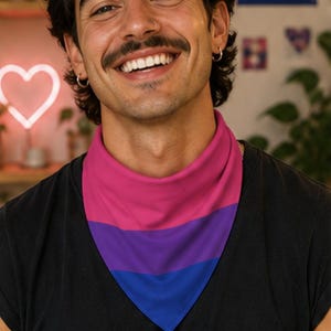 May include: A triangular bandana in the colors of the bisexual pride flag: pink, purple, and blue. The bandana is worn around the neck. The person in the image is wearing a black t-shirt. A sign that says "BI PROUD" is in the background.