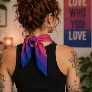 May include: A neck scarf in the colors of the bisexual pride flag, tied in a bow. The scarf is blue, pink, and purple. The person is wearing a black tank top. A poster in the background reads "LOVE WHO YOU LOVE."