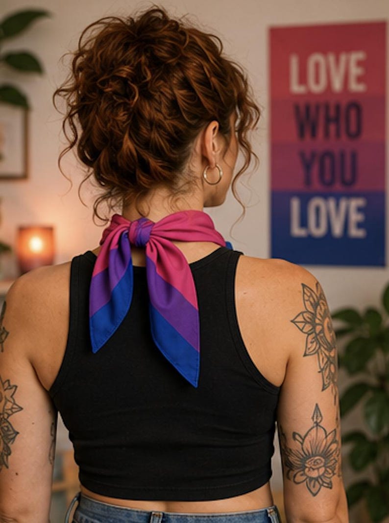 May include: A neck scarf in the colors of the bisexual pride flag, tied in a bow. The scarf is blue, pink, and purple. The person is wearing a black tank top. A poster in the background reads "LOVE WHO YOU LOVE."
