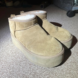 Könnte beinhalten: Ein Paar hellbraune Plateau-Stiefel mit Wildleder-Außenmaterial und einem cremefarbenen, plüschigen Innenfutter. Die Stiefel haben eine runde Spitze und eine dicke, hellbraune Sohle. Die Stiefel sind auf Komfort und Wärme ausgelegt.