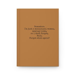 May include: A hardcover notebook with a brown cover. The notebook has the text "Remember. I'm just a menopausal woman, making lists, so I don't forget, Wait. Forget what again?" printed in black.
