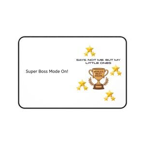 May include: A white rectangular mousepad with a black border. The mousepad features text that reads "Super Boss Mode On!" and "SAYS. NOT ME, BUT MY LITTLE ONES." A trophy with the words "WORLD'S BEST MOM" is also displayed, along with gold stars.