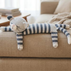 Pode incluir: Um brinquedo de pelúcia de gato de crochê, com listras cinza e azul, deitado em um sofá bege. O gato tem um rosto branco com detalhes em rosa e olhos fechados, e suas patas estão estendidas. O corpo do gato é listrado em cinza e azul.