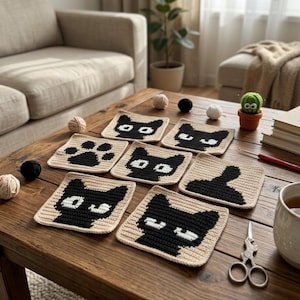 Op de afbeelding: Een verzameling handgemaakte gehaakte onderzetters met een kattenmotief. De onderzetters tonen zwarte kattengezichten en pootafdrukken op een beige achtergrond. Andere items op de tafel zijn bollen wol, een kleine potplant en een kopje thee.