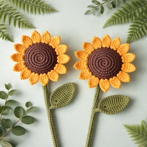 Op de afbeelding: Twee handgemaakte gehaakte zonnebloemen met gele bloemblaadjes en bruine centra. De stelen en bladeren zijn groen. De bloemen zijn gerangschikt op een lichtblauwe achtergrond met groene varenbladeren.