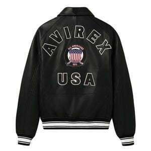Op de afbeelding: Zwarte leren jas met witte letters "AVIREX" en "USA". Een cirkelvormige patch toont de Amerikaanse vlag en de woorden "ALL AMERICANS ESTABLISHED 1975". De jas heeft een kraag, geribde manchetten en een gestreepte zoom.