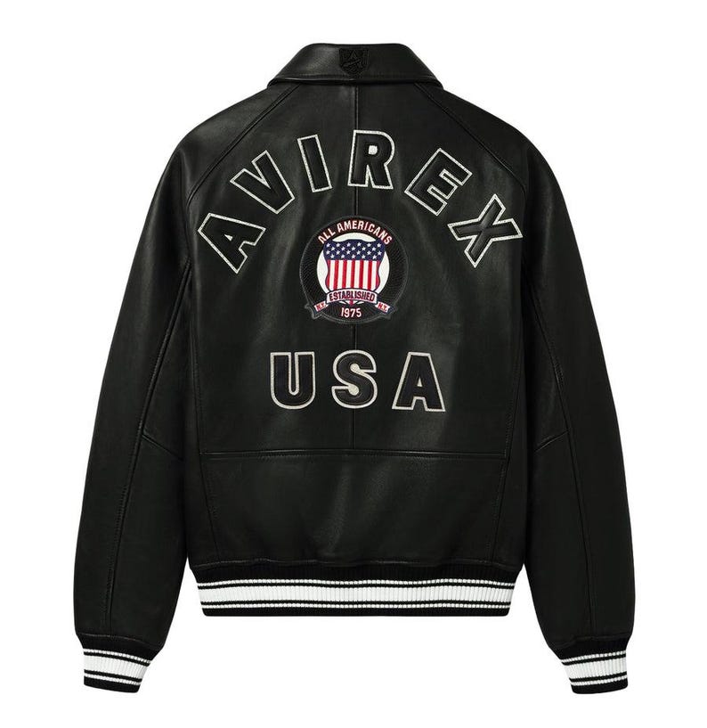 Op de afbeelding: Zwarte leren jas met witte letters "AVIREX" en "USA". Een cirkelvormige patch toont de Amerikaanse vlag en de woorden "ALL AMERICANS ESTABLISHED 1975". De jas heeft een kraag, geribde manchetten en een gestreepte zoom.