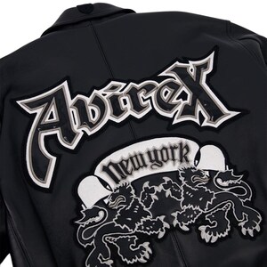K&ouml;nnte beinhalten: Schwarze Lederjacke mit dem Avirex-Logo in Wei&szlig; und Grau, einem New York-Banner und zwei L&ouml;wenemblemen. Die Jacke hat einen klassischen Kragen und einen Knopf oben.