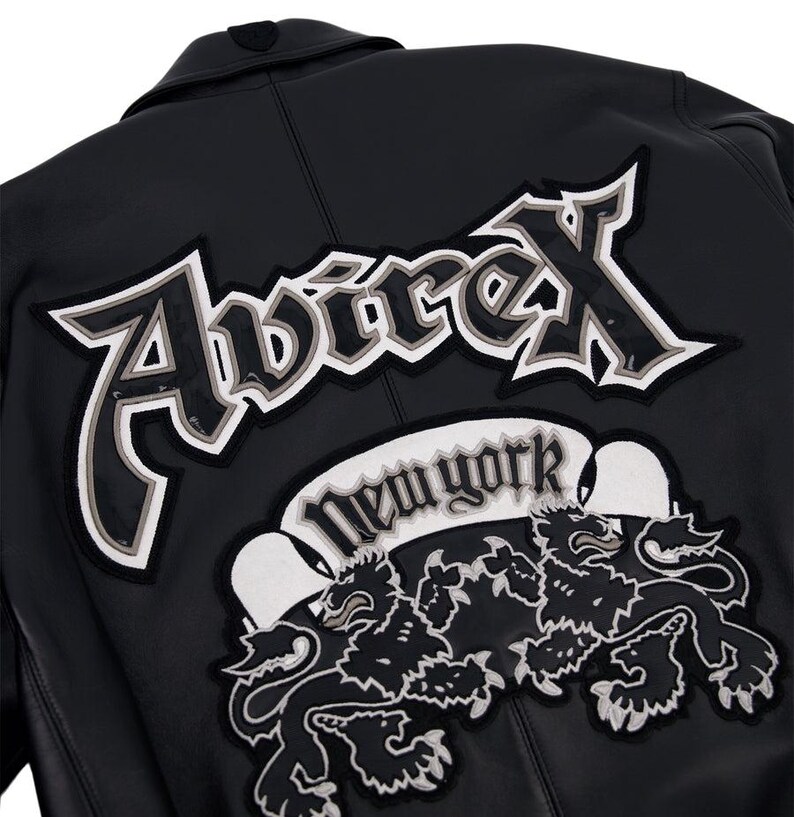 K&ouml;nnte beinhalten: Schwarze Lederjacke mit dem Avirex-Logo in Wei&szlig; und Grau, einem New York-Banner und zwei L&ouml;wenemblemen. Die Jacke hat einen klassischen Kragen und einen Knopf oben.