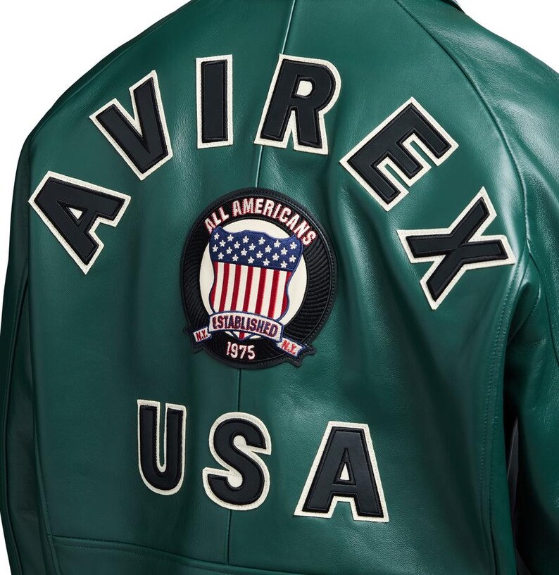May include: A green leather jacket with white and black lettering and patches. The back of the jacket features the word "AVIREX" across the top, "USA" at the bottom, and a circular patch with an American flag design.