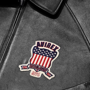 Op de afbeelding: Close-up van een zwarte leren jas met een decoratieve patch. De patch heeft een schildontwerp met rode en witte strepen, blauwe sterren en de tekst "AVIREX NY ESTABLISHED 1975". De jas heeft een rits en een kraag.