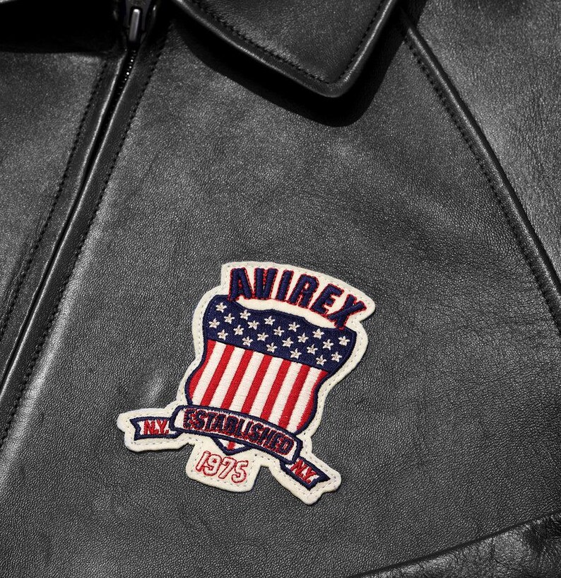 Op de afbeelding: Close-up van een zwarte leren jas met een decoratieve patch. De patch heeft een schildontwerp met rode en witte strepen, blauwe sterren en de tekst "AVIREX NY ESTABLISHED 1975". De jas heeft een rits en een kraag.