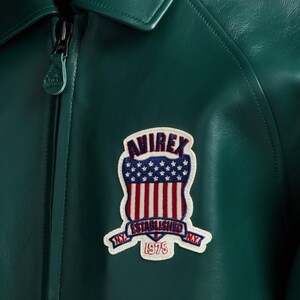 May include: Close-up of a dark green leather jacket with a zipper closure. The jacket features a patch with the text "AVIREX ESTABLISHED 1975 N.Y." in red, white, and blue. The patch is in the shape of a shield with stars and stripes.