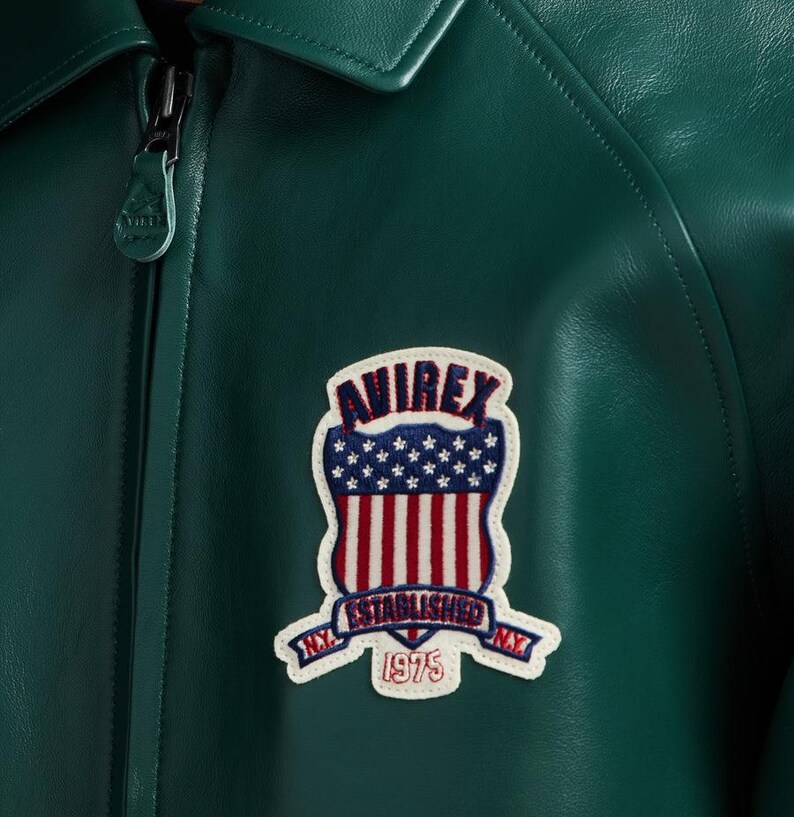 May include: Close-up of a dark green leather jacket with a zipper closure. The jacket features a patch with the text "AVIREX ESTABLISHED 1975 N.Y." in red, white, and blue. The patch is in the shape of a shield with stars and stripes.