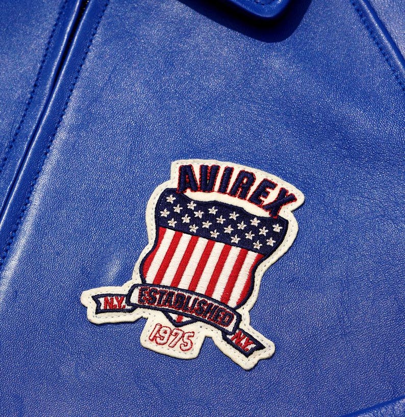 May include: Close-up of a blue leather jacket with a patch. The patch features the word "AVIREX" above a shield design with red and white stripes and stars. The patch also includes the text "ESTABLISHED 1975" and "NY".
