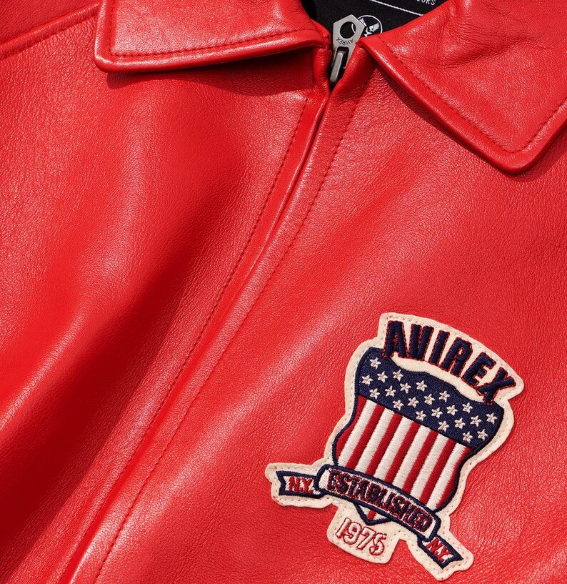 Peut inclure: Gros plan sur une veste en cuir rouge avec un col. La veste pr&eacute;sente un &eacute;cusson avec le texte "AVIREX" au-dessus d'un motif de bouclier avec le drapeau am&eacute;ricain et le texte "NY ESTABLISHED 1975".