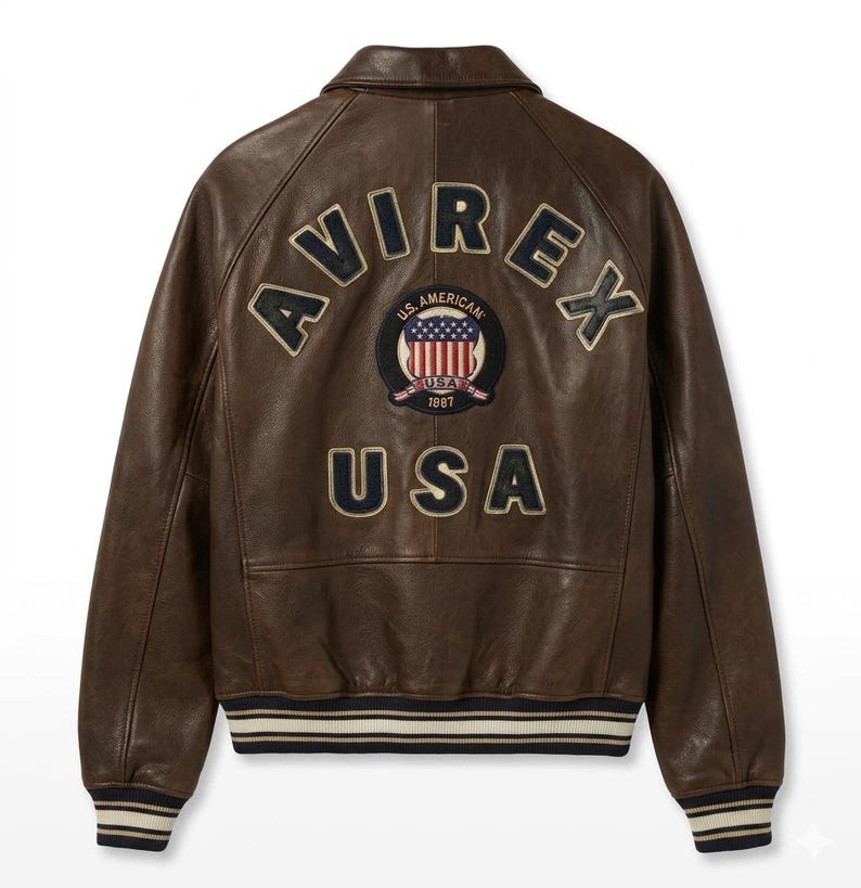 Op de afbeelding: Een bruine leren jas met het woord "AVIREX" over de bovenrug. Daaronder een patch met de woorden "U.S. AMERICAN" en "USA 1987" rond een afbeelding van de Amerikaanse vlag. Het woord "USA" staat eronder.