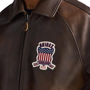 Op de afbeelding: Bruine leren jas met ritssluiting en kraag. De jas heeft een patch met de tekst "AVIREX ESTABLISHED NY 1975" in een schildontwerp met rode en witte strepen en blauwe sterren.