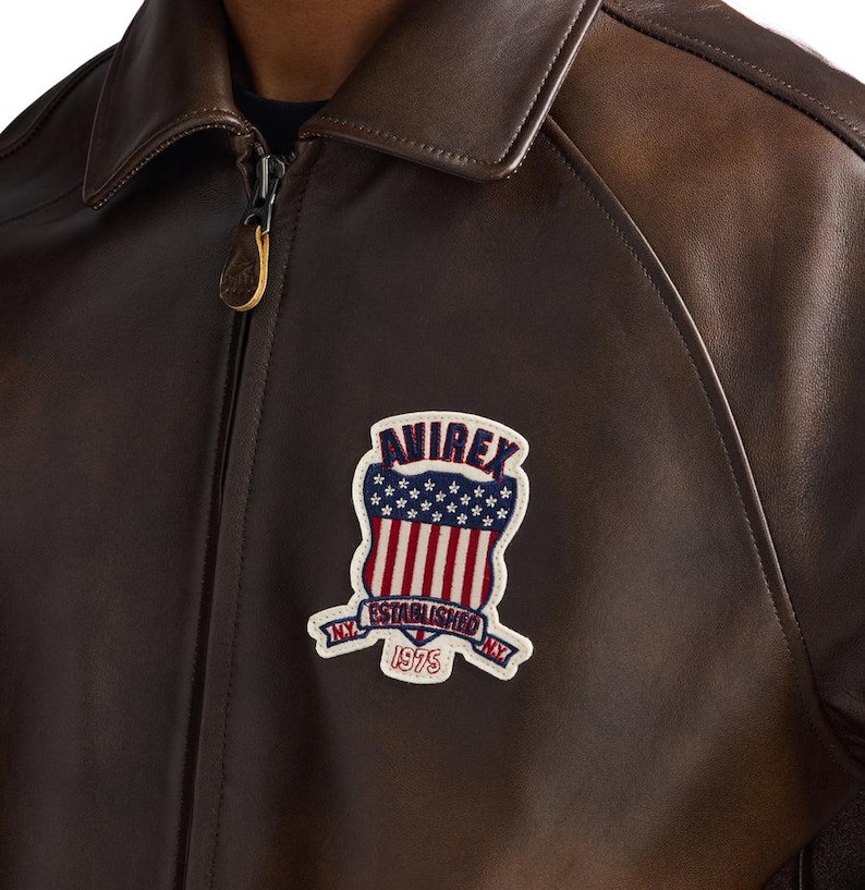 Op de afbeelding: Bruine leren jas met ritssluiting en kraag. De jas heeft een patch met de tekst "AVIREX ESTABLISHED NY 1975" in een schildontwerp met rode en witte strepen en blauwe sterren.